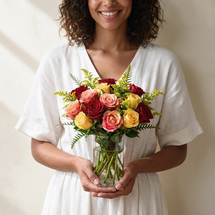 Small size: 18 red, peach and yellow roses in a small glass vase held by a smiling woman with curly hair, Fabulous Flowers South Africa