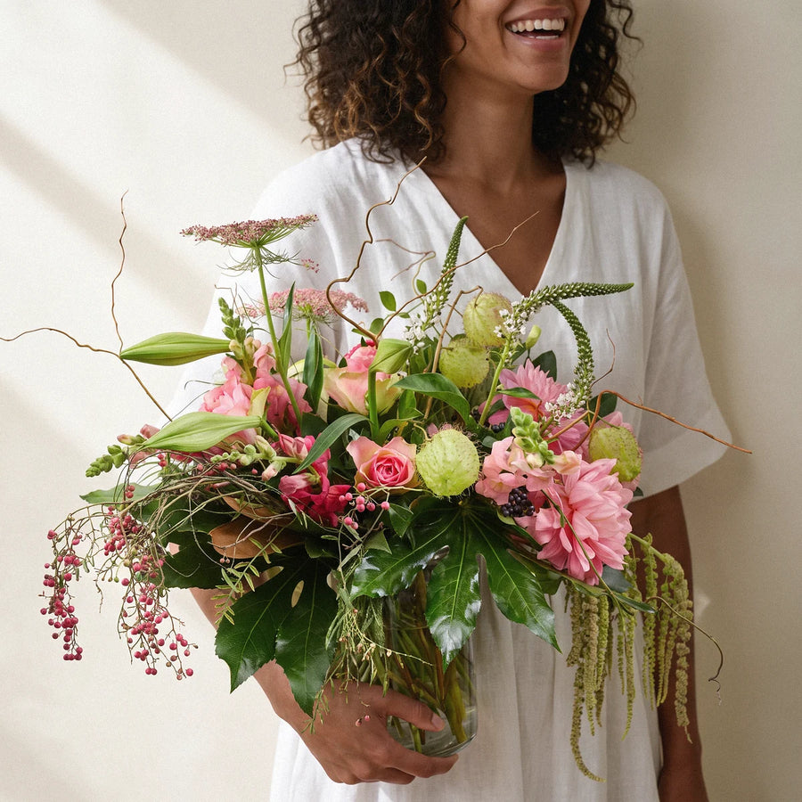 Laughing woman with curly hair in a white linen dress carrying a wild pink garden-style vase of flowers on her birthday, Fabulous Flowers South Africa