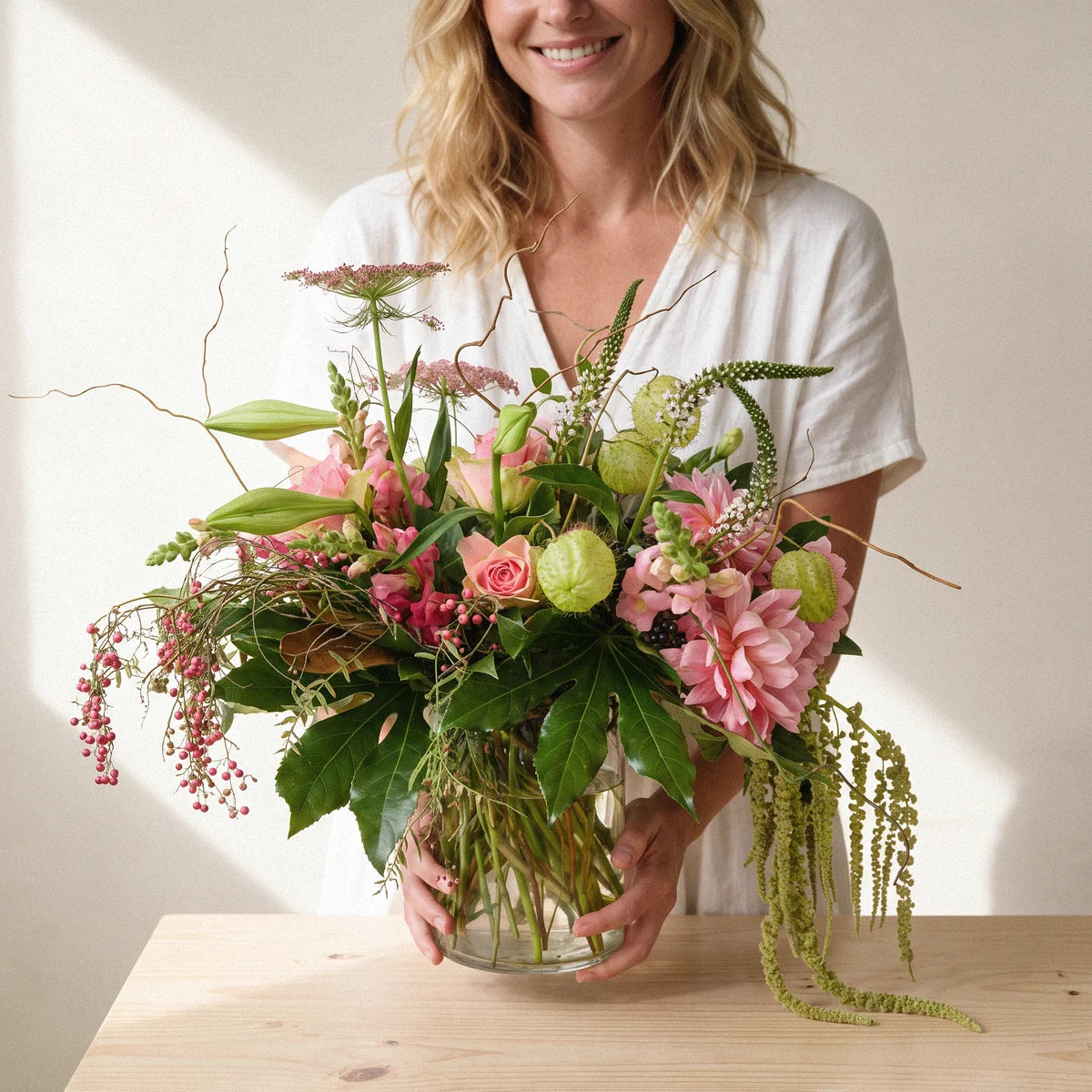 Woman in a white linen dress setting pink and green flowers in a glass vase down on a pine table, Fabulous Flowers South Africa