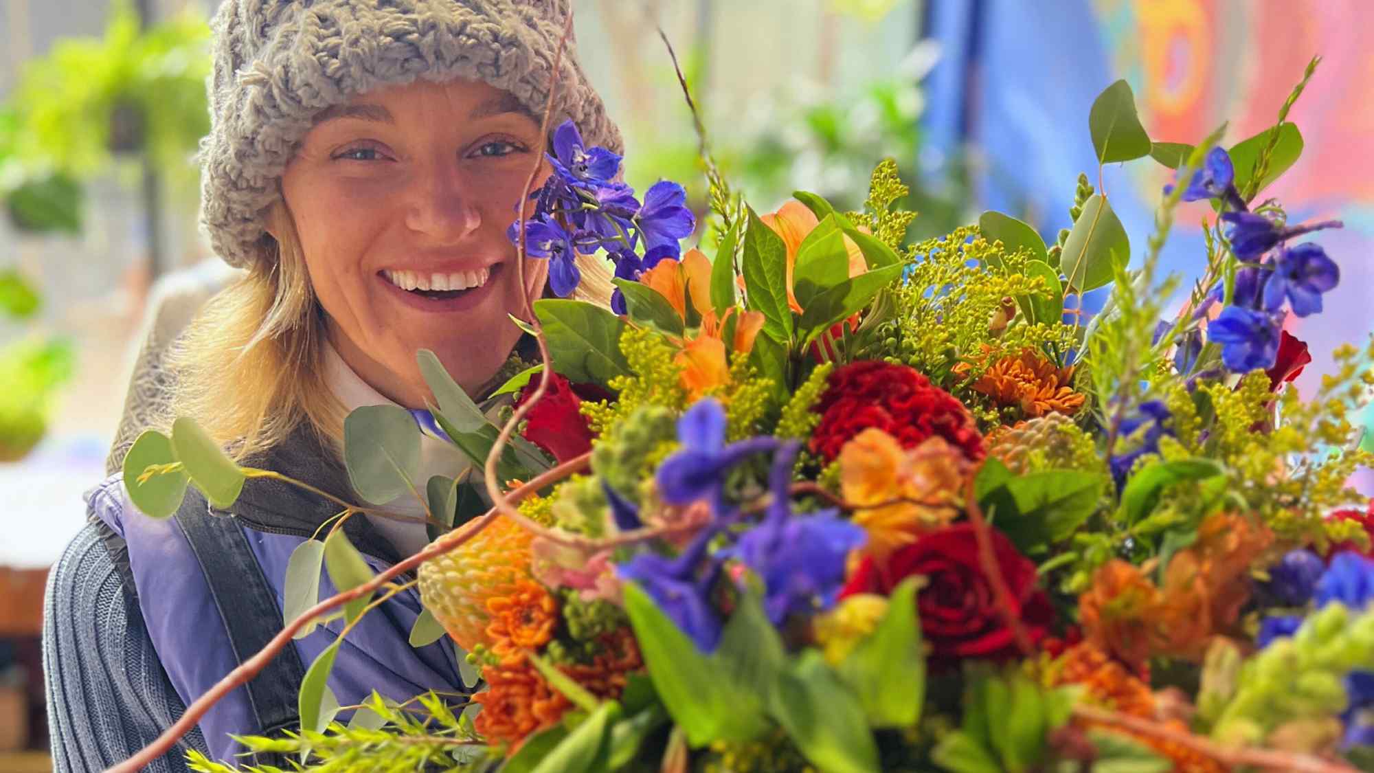 A smiling florist in a knit hat holding a vibrant bouquet with orange, red, and blue flowers, showcasing a mix of greenery and colourful blooms.