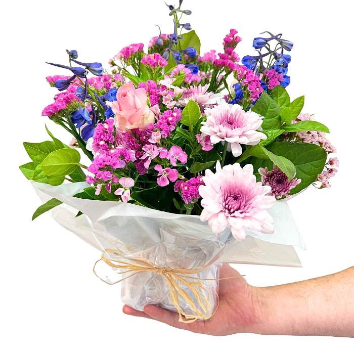 Alchemy Posy hand-tied posy with pink, lilac and blue blooms wrapped in white paper, held in hand