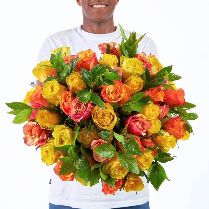 All The Ways We Bloom bouquet with approx. 50 yellow, orange, and coral roses, held by a smiling person in a white t-shirt.