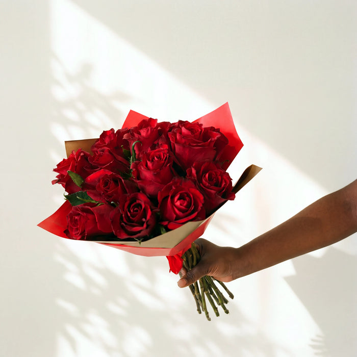 A dozen red roses hand-tied in red and gold wrap, held out by a florist from the Amour bouquet range