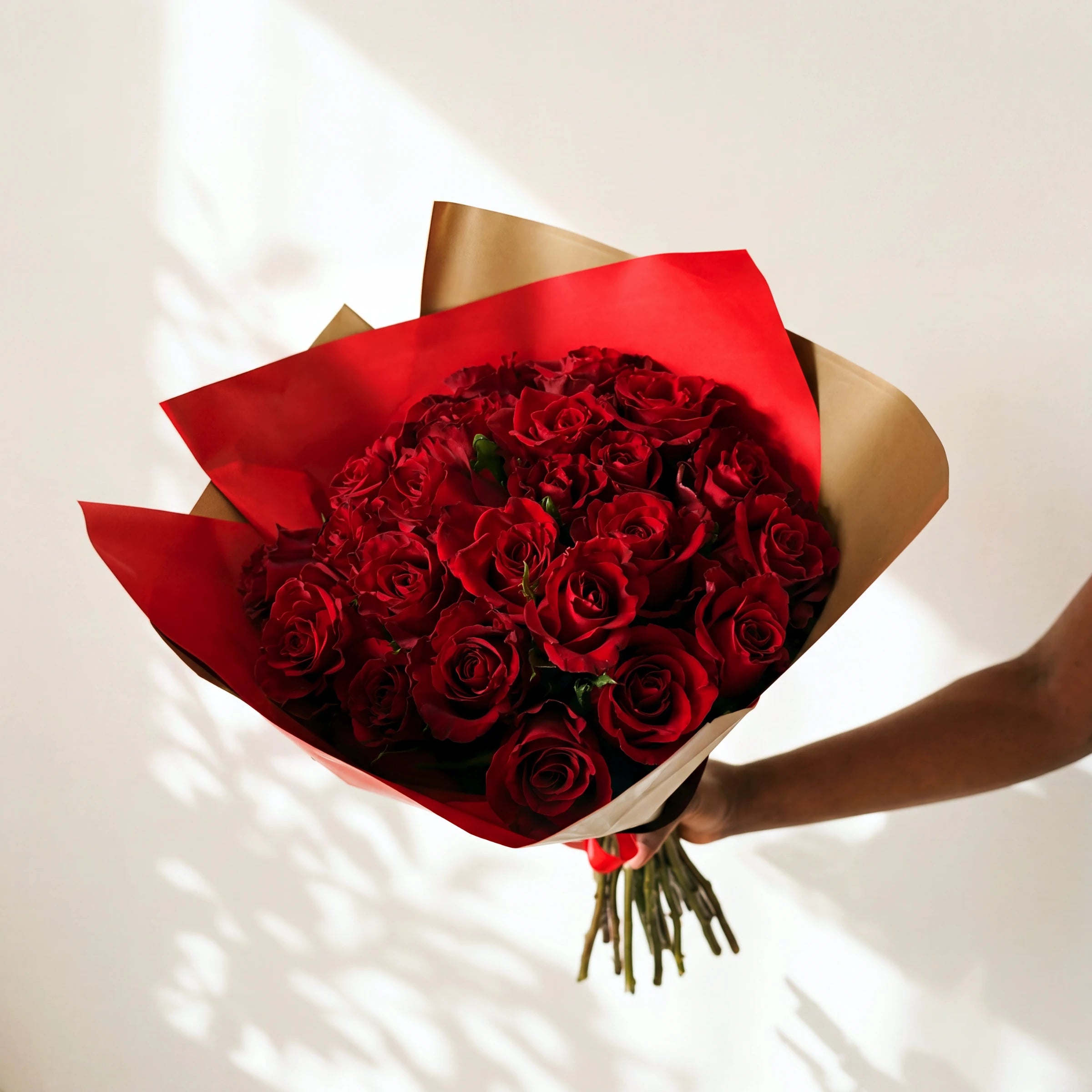 Two dozen red roses in the Amour bouquet, wrapped in red and gold paper against a white wall