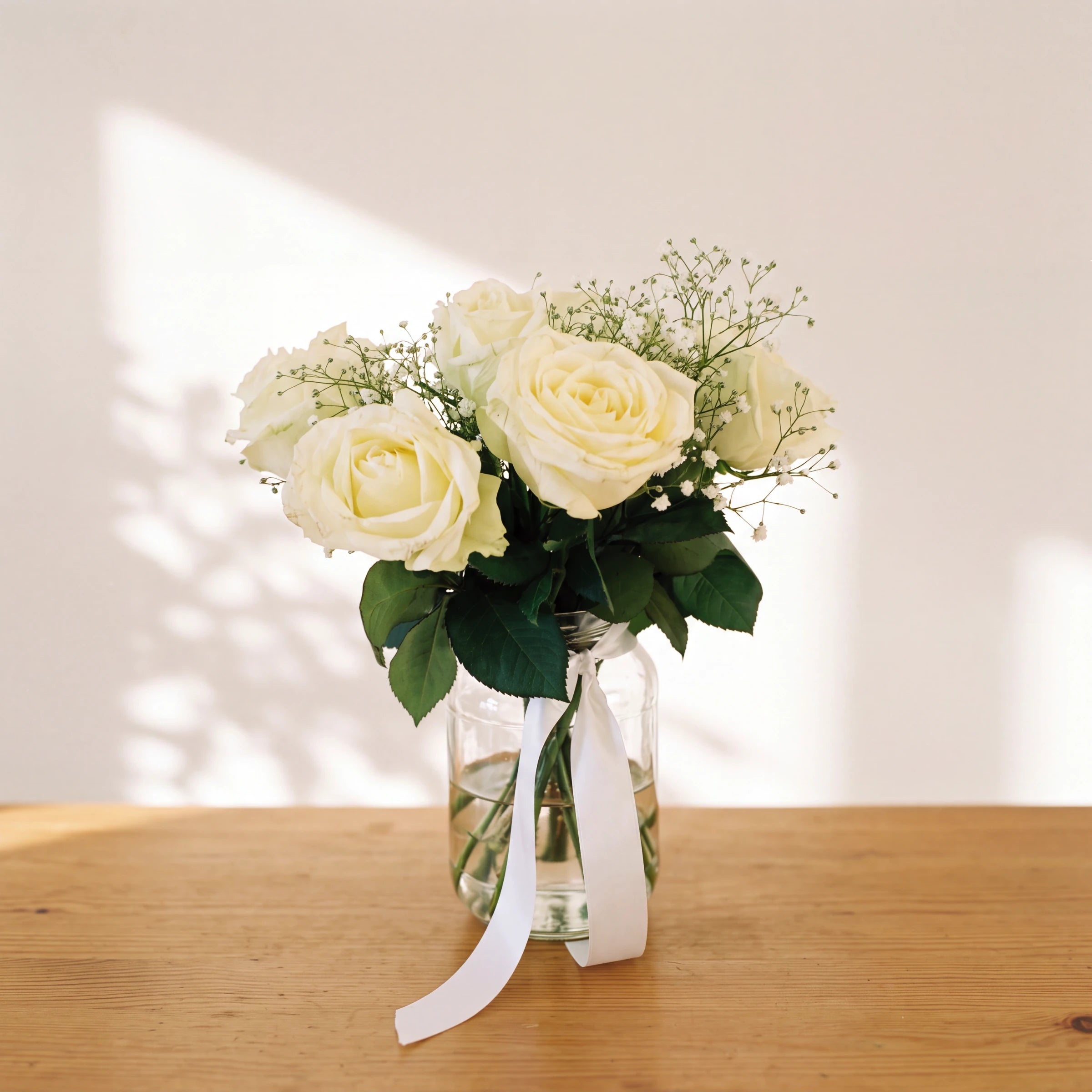Angel White arrangement of ivory white roses and baby's breath in a glass jar with a white ribbon on a wooden table