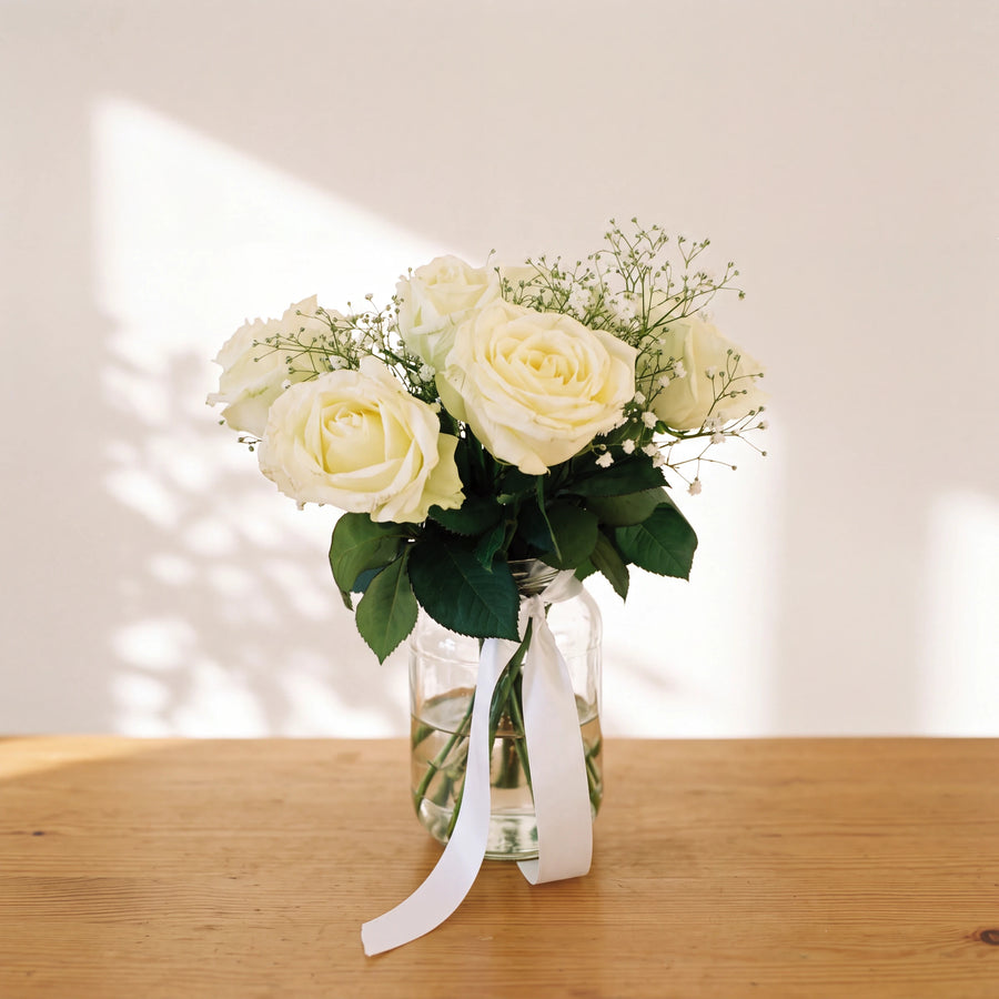 Angel White arrangement of ivory white roses and baby's breath in a glass jar with a white ribbon on a wooden table