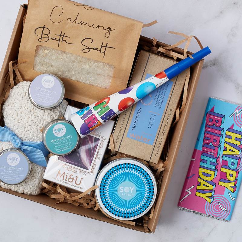 Close-up of the Birthday Treats & Tranquility Gift Box featuring bath salts, soy candles, a birthday party blower, and a 'Happy Birthday' chocolate bar.