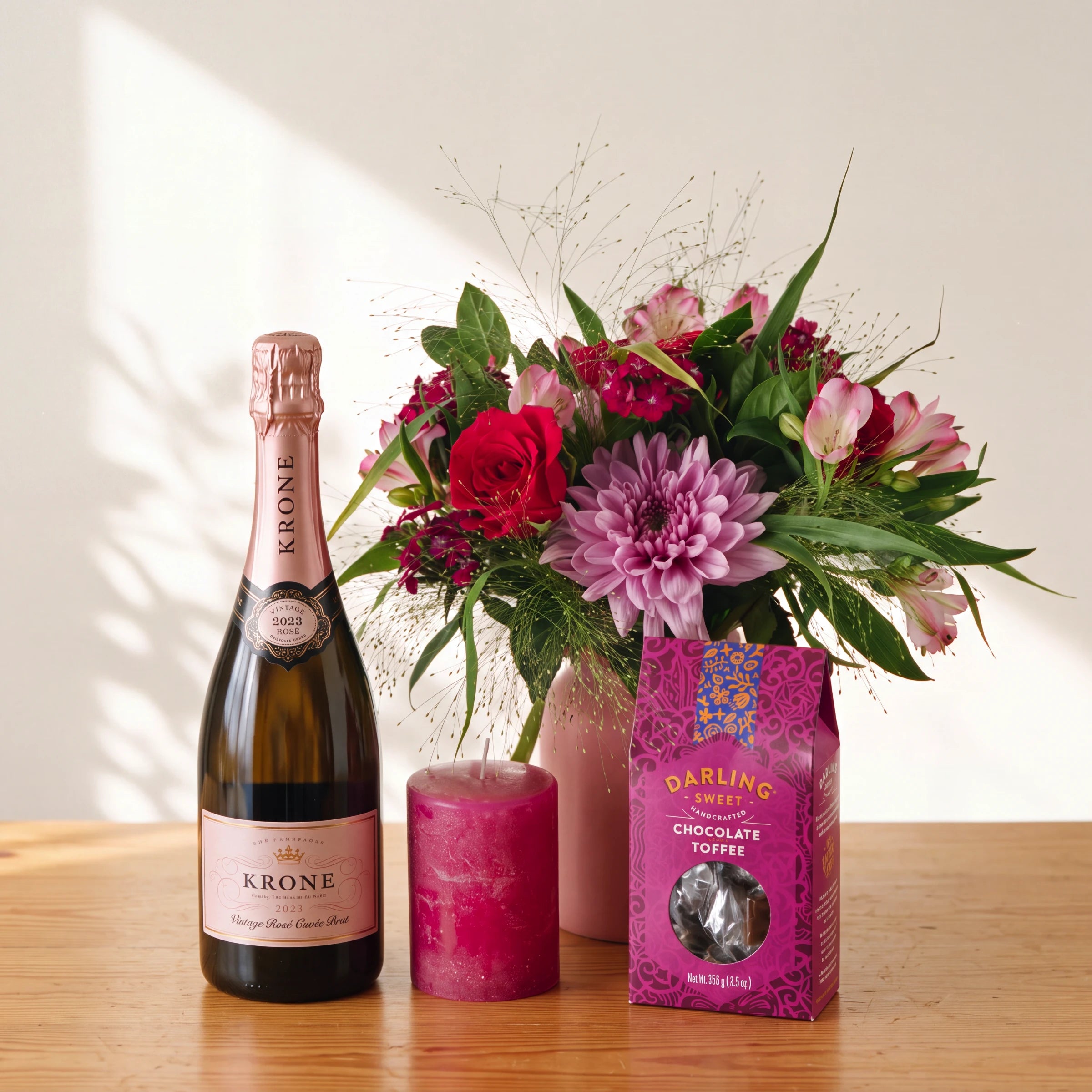 A Krone sparkling wine bottle, a bright pink candle, and a pink box of Darling Cream Chocolate Toffee are arranged on a wooden table beside a pink vase filled with a colorful flower arrangement against a warm off-white wall.