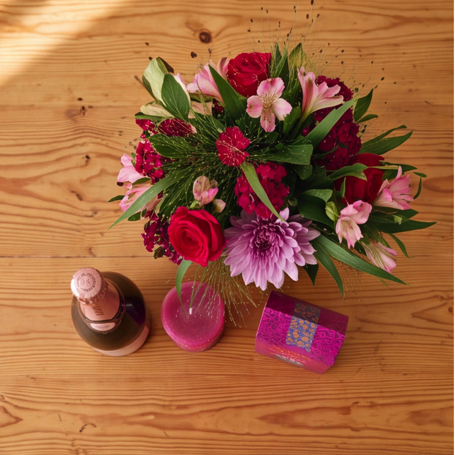 Overhead view of the Bloom and Bubble gift set on a wooden table, a red rose and lilac chrysanthemum jar arrangement beside Krone Rosé Cuvée Brut, a berry-pink candle and Darling Sweet toffee