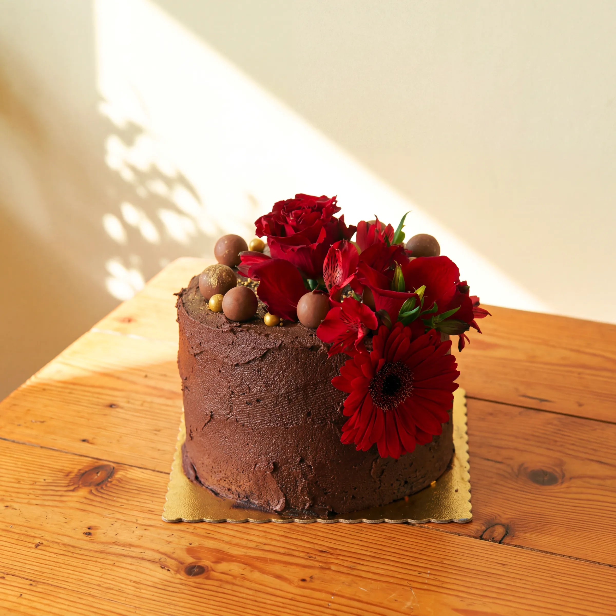 Side angle of the Fabulous Flowers chocolate cake on a gold scalloped board, finished with a fresh red rose and gerbera daisy floral arrangement and chocolate truffle accents
