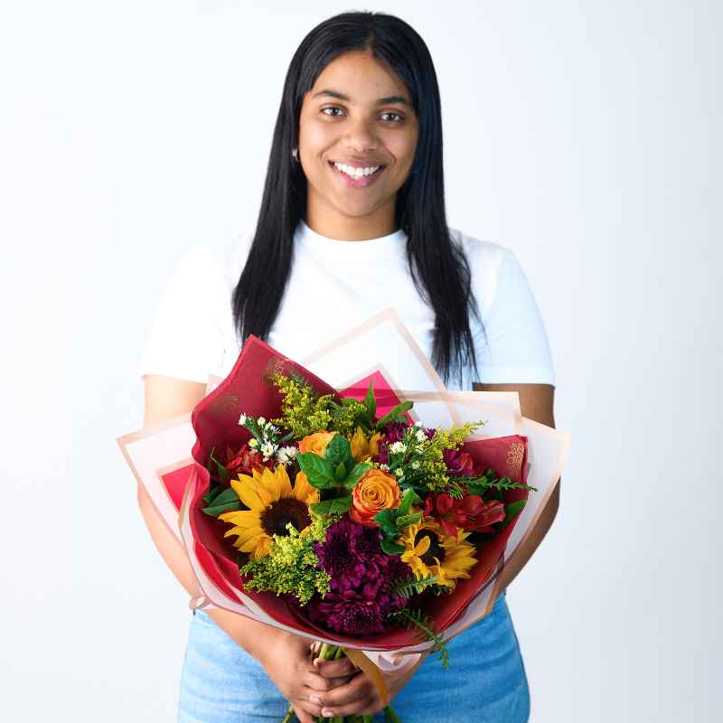 Blooming Sunshine Flower Bouquet - A woman holding a vibrant bouquet of sunflowers, roses, and assorted colourful flowers wrapped in red and pastel paper.