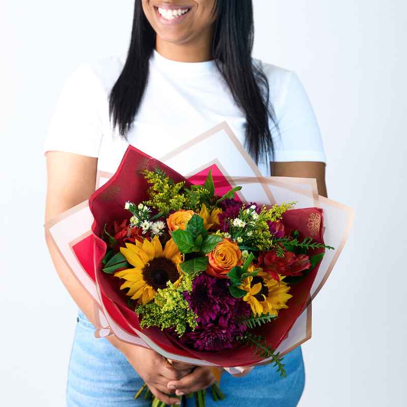 Blooming Sunshine Flower Bouquet - Close-up of a woman holding a bouquet featuring sunflowers, roses, and various colourful flowers wrapped in red and pastel paper.