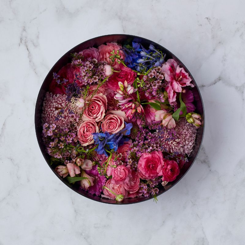 Overhead view of a luxurious round floral arrangement with pink roses, purple delphiniums, ranunculus, and wax flowers.