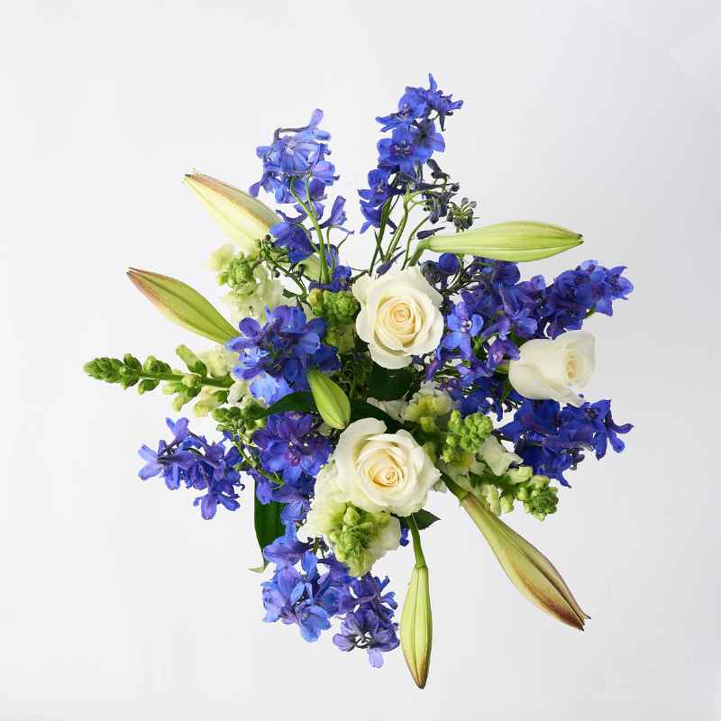 Top view of the Blue Sapphire flower arrangement, showcasing white roses, blue delphiniums, and unopened lilies in a clear glass jar.