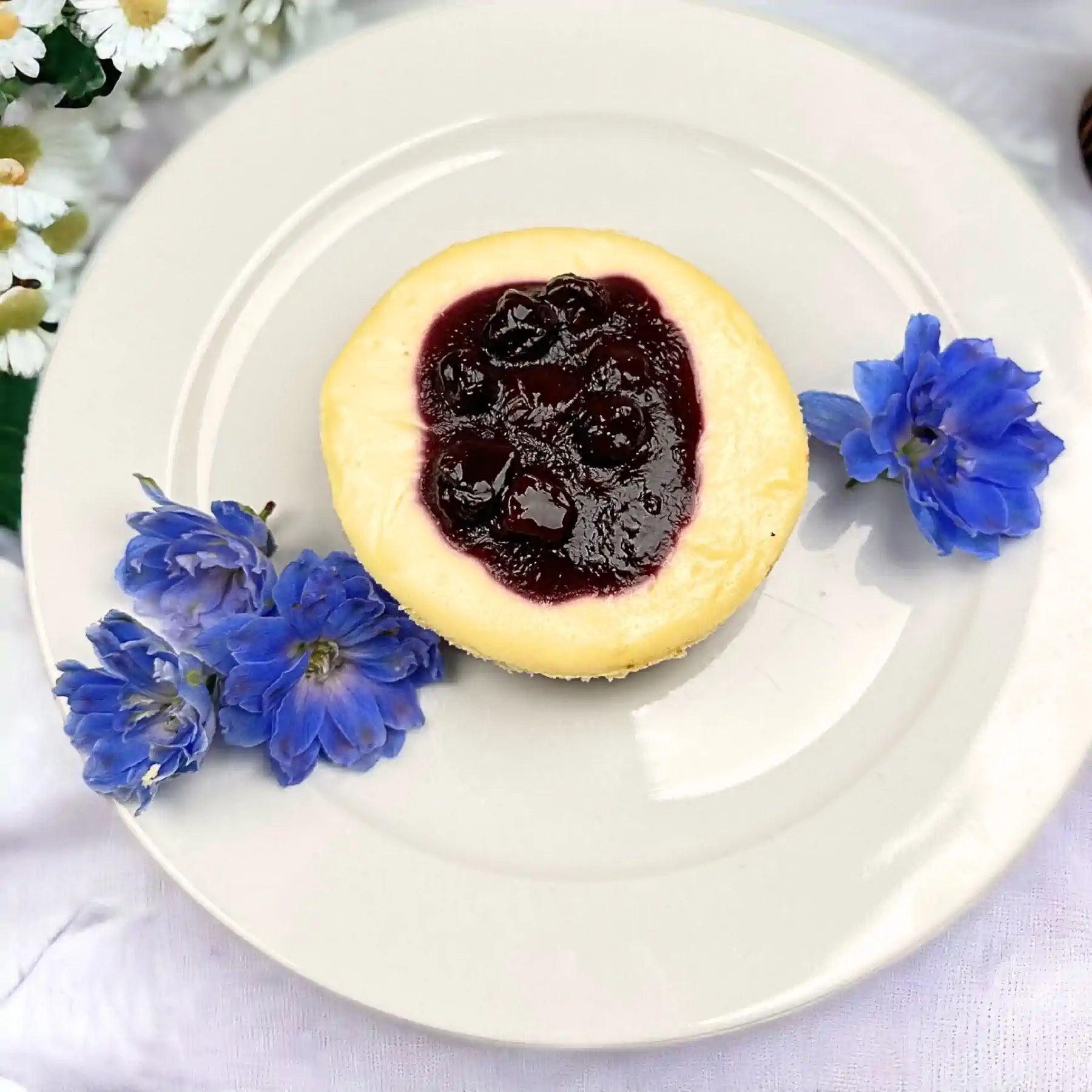 Elegant blueberry cheesecake with a luscious blueberry topping, complemented by blue flowers on a white plate. Fabulous Flowers and Gifts.