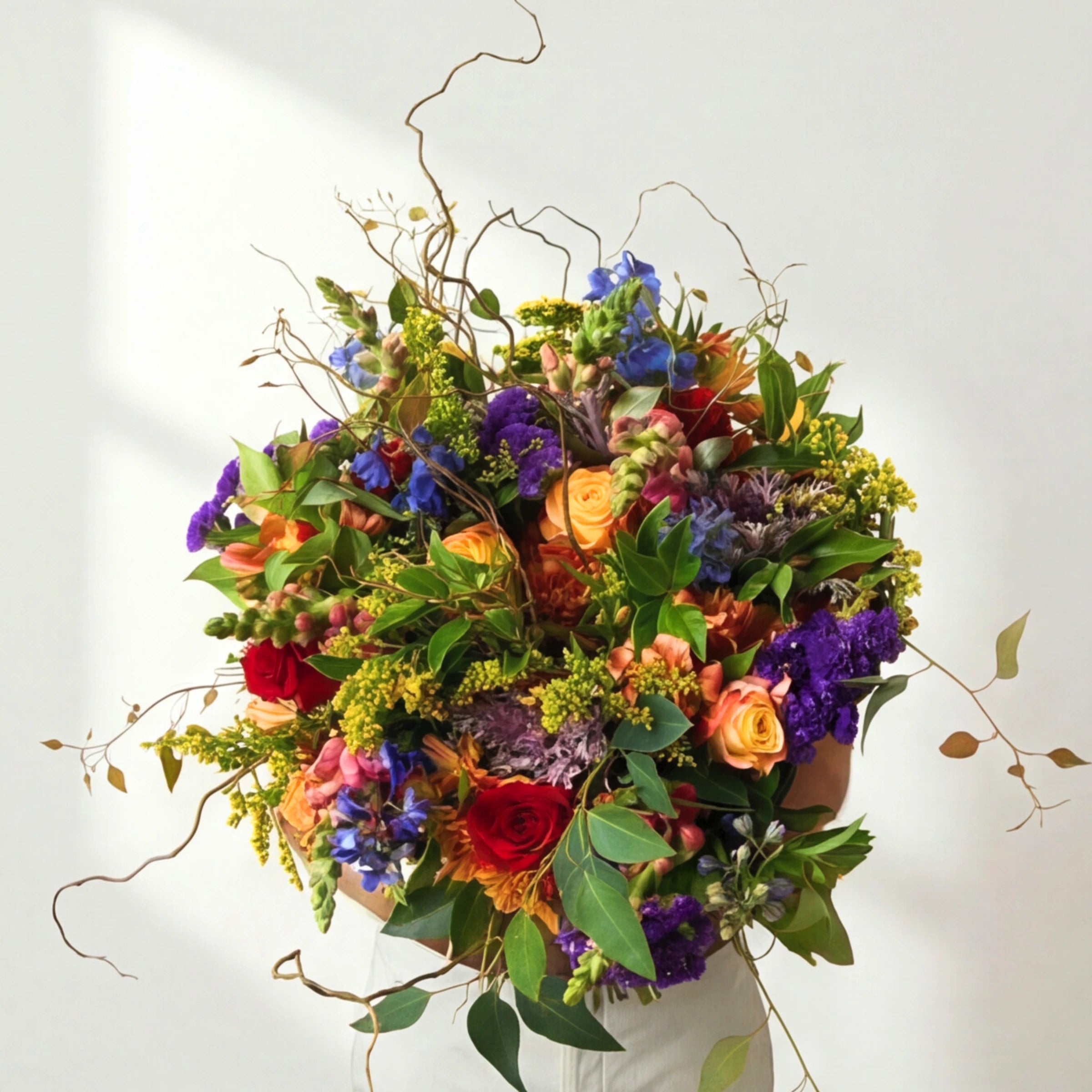 Brilliantly Happy Bouquet held by a man in white, showing orange roses, blue delphinium, purple statice, ornamental kale, solidago and curly willow branches against a soft white background, Fabulous Flowers Cape Town