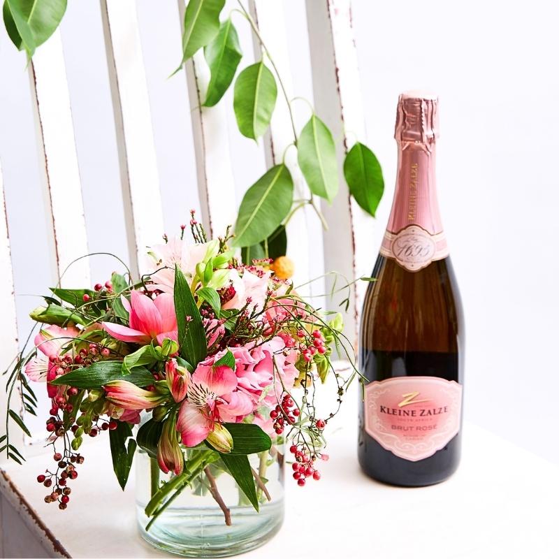 Pink flower arrangement in glass vase beside sparkling wine bottle on a bright windowsill