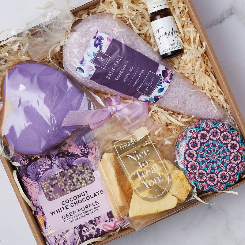 Close-up of Calm and Comfort Gift Box with purple bath bomb, lavender bath salts, coconut white chocolate, treats, and scented oil for relaxation.
