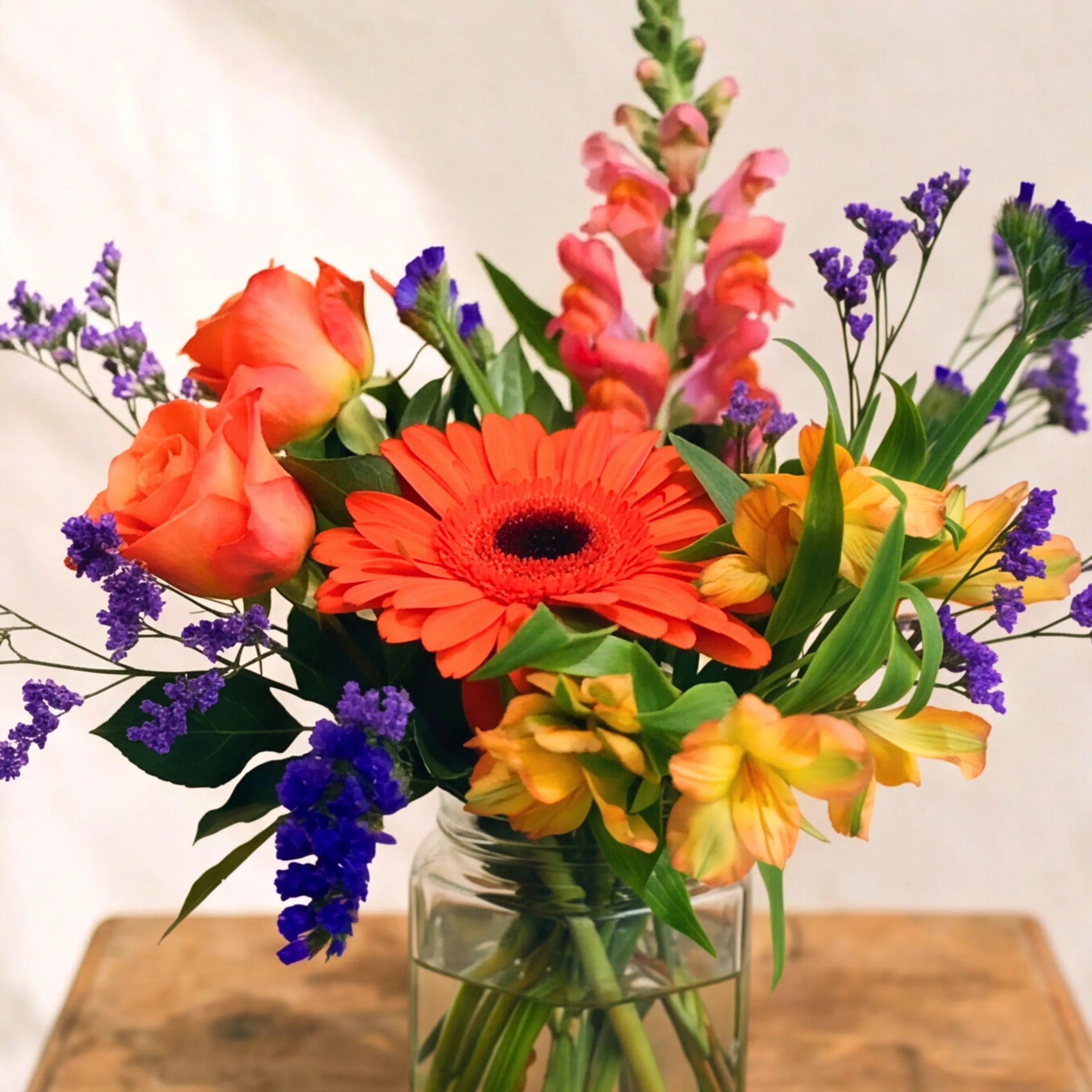 Top-down view of Carnival in a Jar from Fabulous Flowers South Africa, an orange gerbera daisy at the centre with peach roses, pink snapdragons, yellow alstroemeria and purple statice in a small glass jar, the affordable mixed flower bouquet in our flowers-in-a-jar series