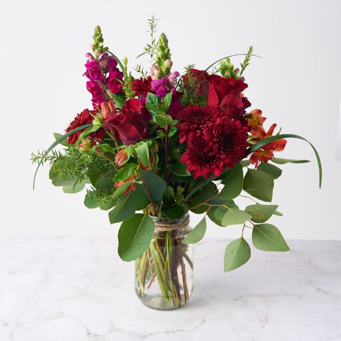 A bouquet in a glass jar featuring red roses, gerbera daisies, snapdragons, and lush greenery on a white background.