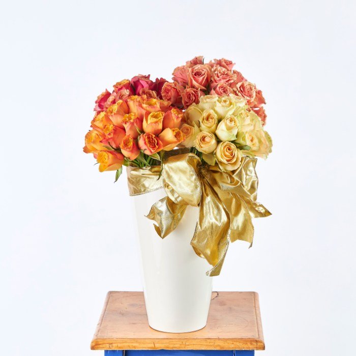 Cherry Elegance flower arrangement in a white vase with golden ribbon, showcasing layers of pink, yellow, and orange roses.