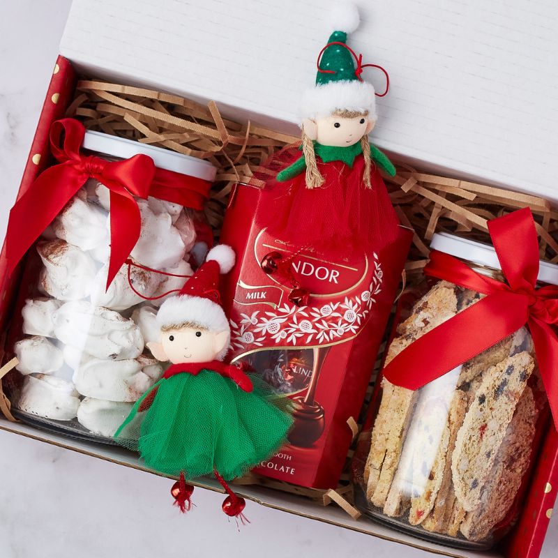 Close-up of festive gift box containing Lindor chocolate, meringue cookies, biscotti, and two elf dolls with red and green outfits.