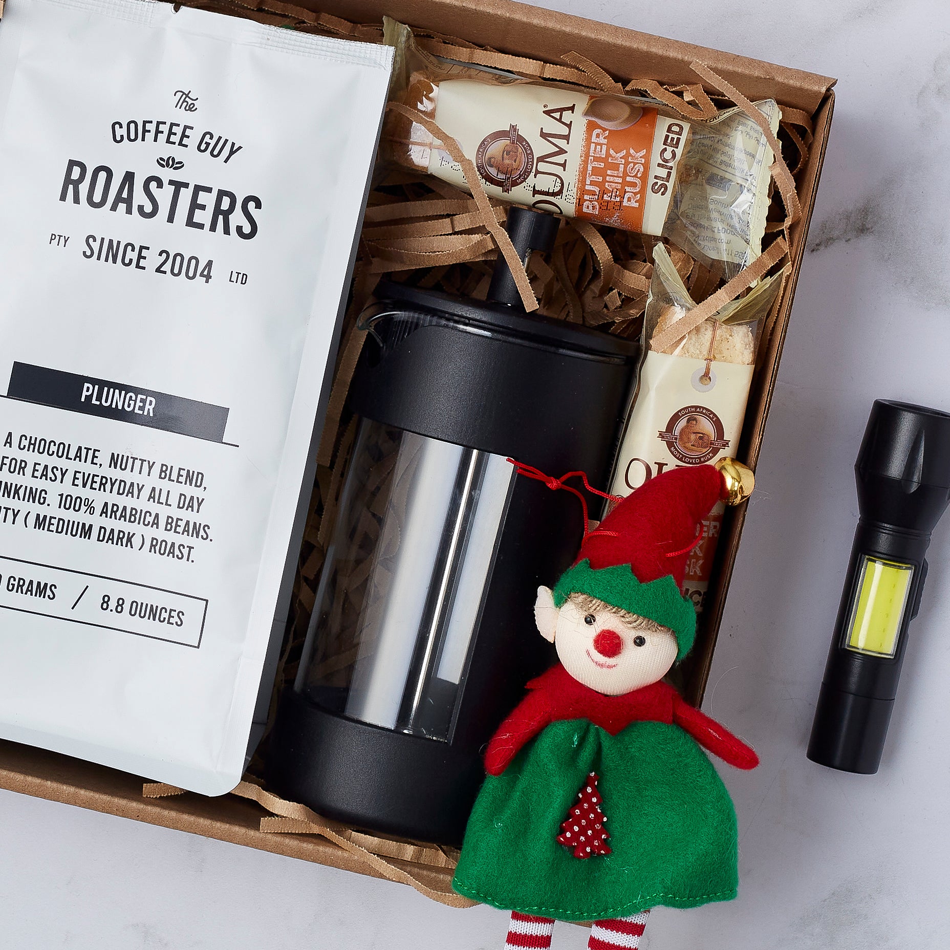 Festive coffee gift box with plunger coffee, French press, Ouma rusks, elf decoration, and mini LED torch, perfect for Christmas.
