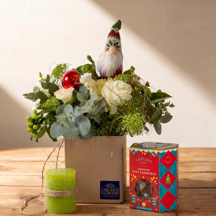 White roses, eucalyptus and a knitted Christmas gnome in a kraft gift box, with a lime green candle and a box of soft caramels on a wooden table