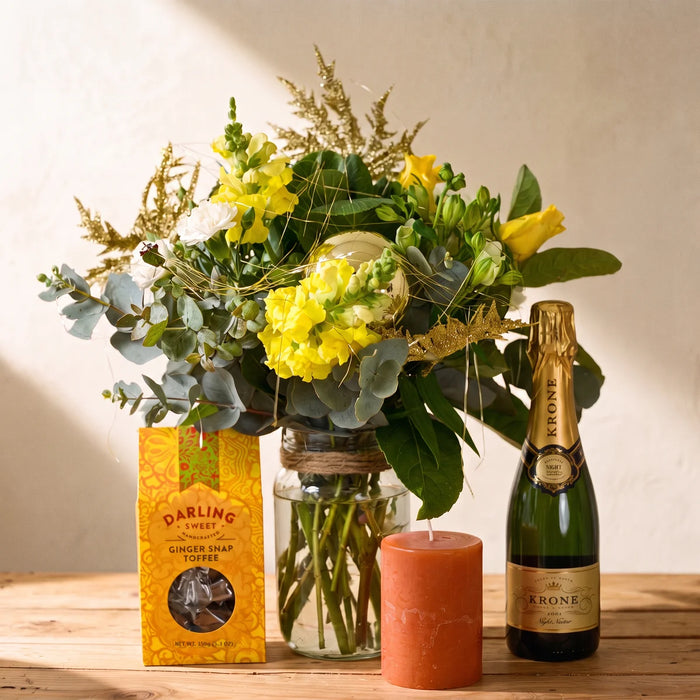 Glass jar of yellow snapdragons, eucalyptus and gold sprays tied with twine, beside a half bottle of Krone Night Nectar, ginger snap toffee and an orange candle, Fabulous Flowers South Africa
