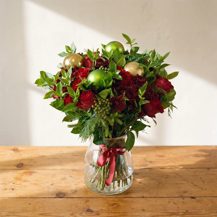 Christmas bouquet of red roses, burgundy chrysanthemums and gold and green baubles in a glass vase on a pine table, Fabulous Flowers South Africa