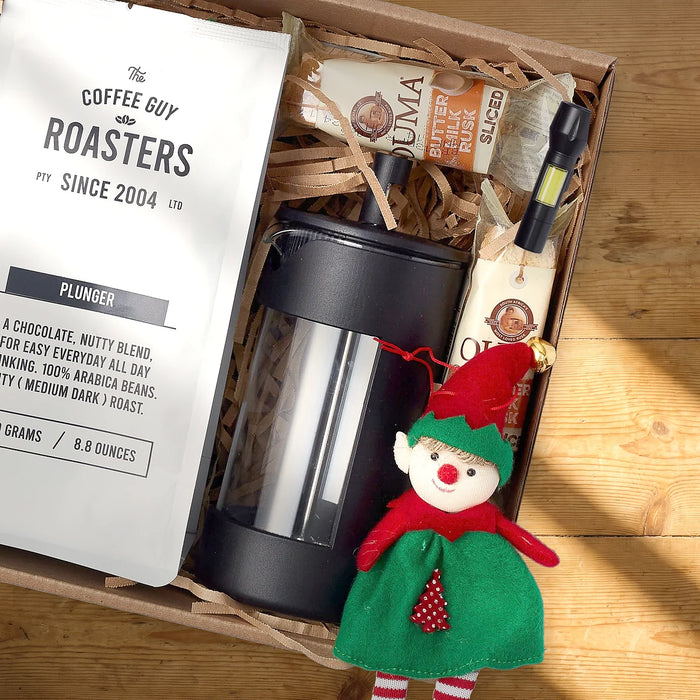 Open kraft gift box from above with a bag of The Coffee Guy Roasters plunger coffee, a black French press, two packs of Ouma buttermilk rusks, a small LED torch and a felt Christmas elf on a sunlit pine table, Fabulous Flowers South Africa