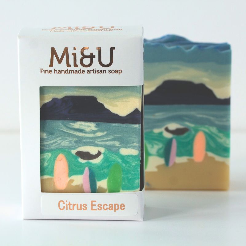 Handcrafted Citrus Escape Soap by Mi&U with ocean artwork and pastel surfboards on a golden sandy shore.