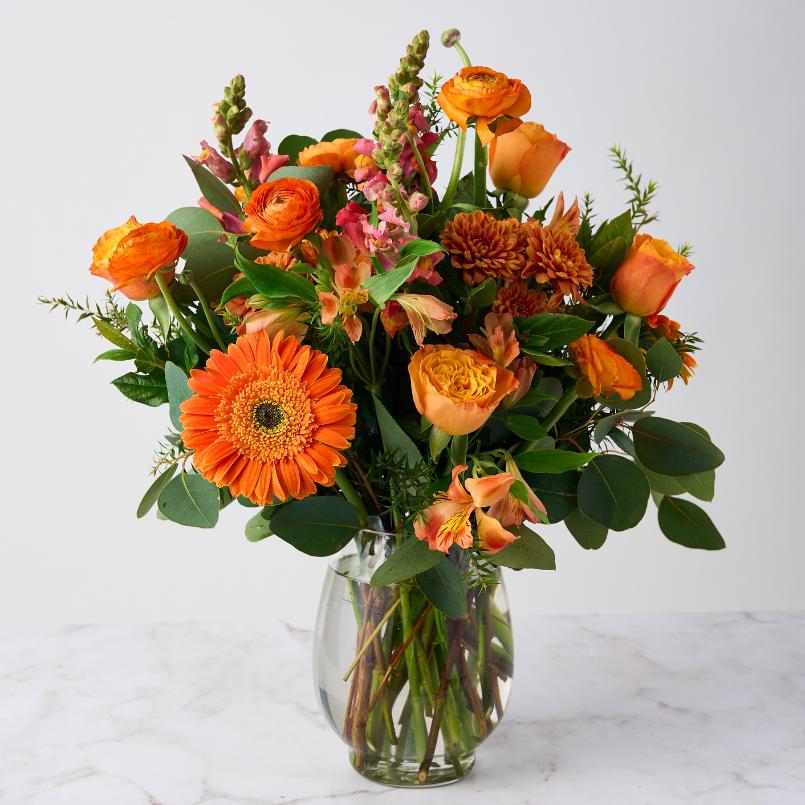 Citrus Couture Flower Arrangement in a clear glass vase, featuring vibrant orange gerberas, roses, snapdragons, and lush greenery.
