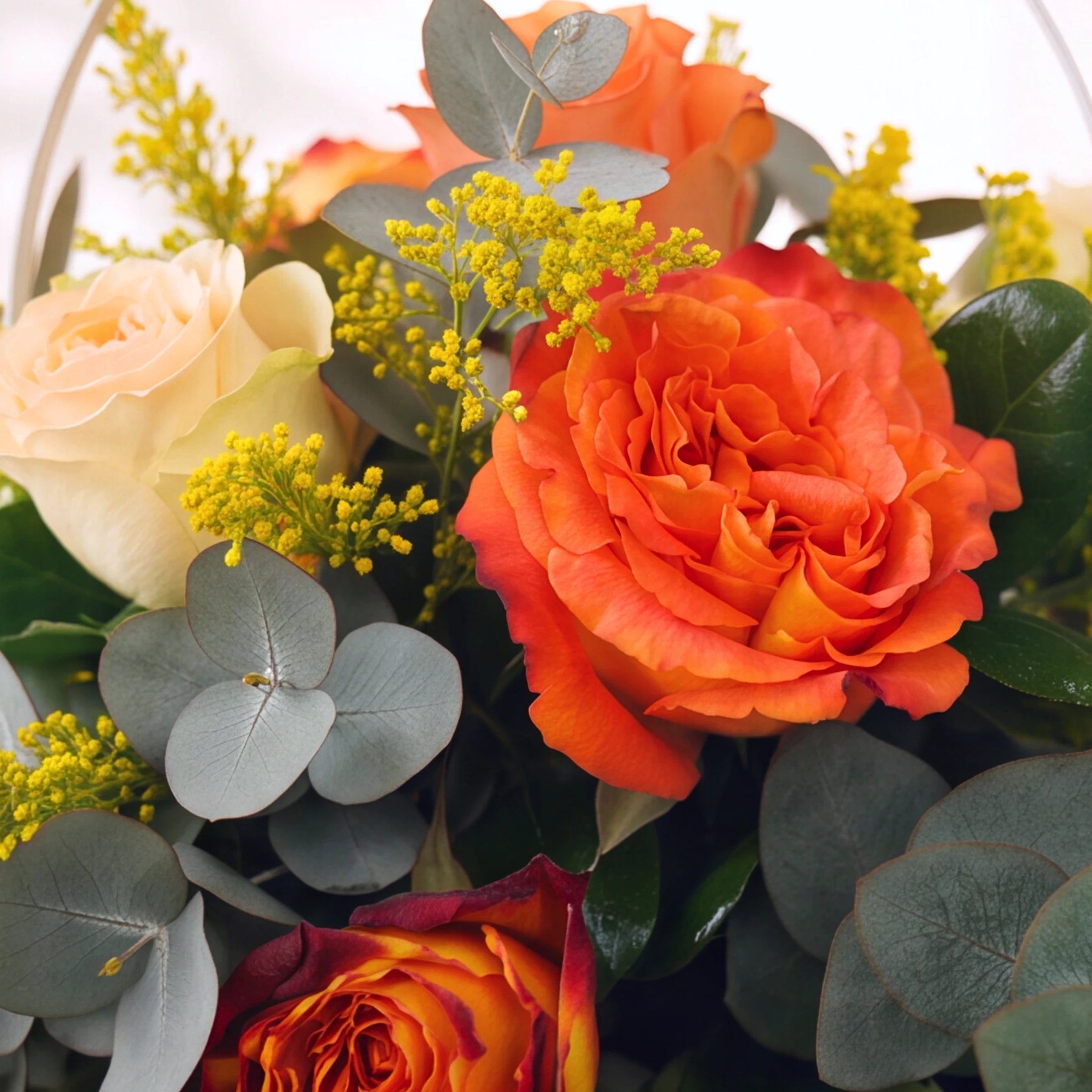 A close-up of a floral arrangement featuring vibrant orange roses, pale peach carnations, red and yellow marigold roses, and dusty blue-green eucalyptus leaves with small yellow filler flowers.