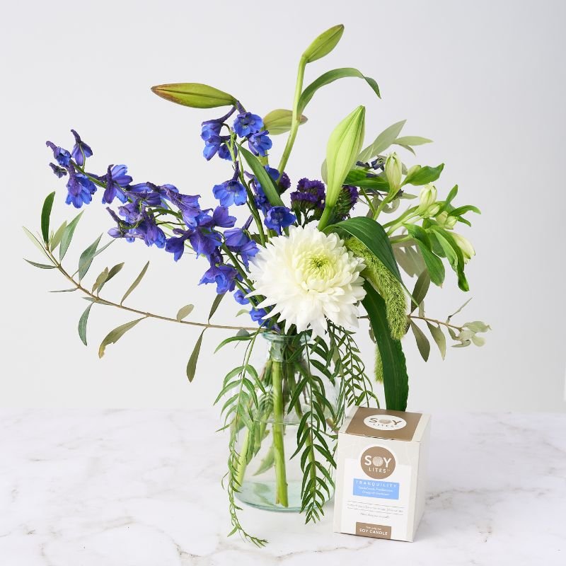 Elegant arrangement of blue delphiniums, white chrysanthemum, and lush greenery in a glass vase, paired with a soothing Soy Lites Tranquility scented candle.