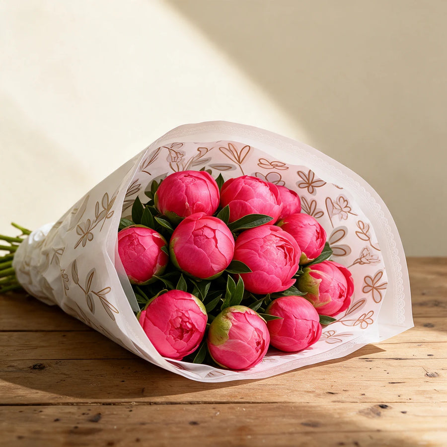 Wrapped bouquet of coral peony buds with green leaves resting on a wooden table in soft window light
