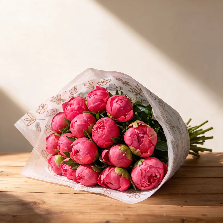 Wrapped armful of coral peony buds with green leaves resting on a wooden table, stems showing at the back