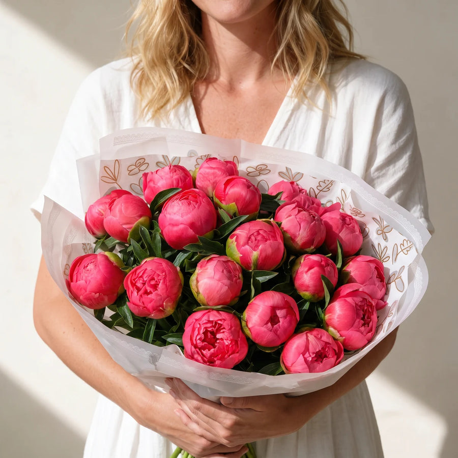 Woman with blonde hair holding a full wrapped bouquet of coral peonies with both arms