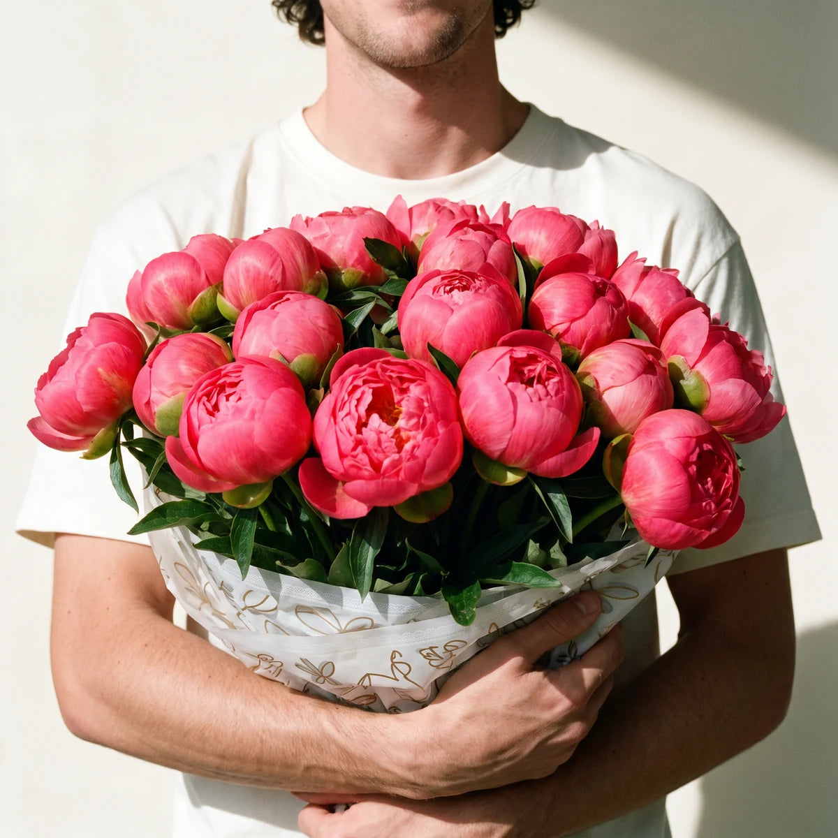 Man in a white T-shirt holding a big rounded bouquet of coral peonies against a sunlit wall