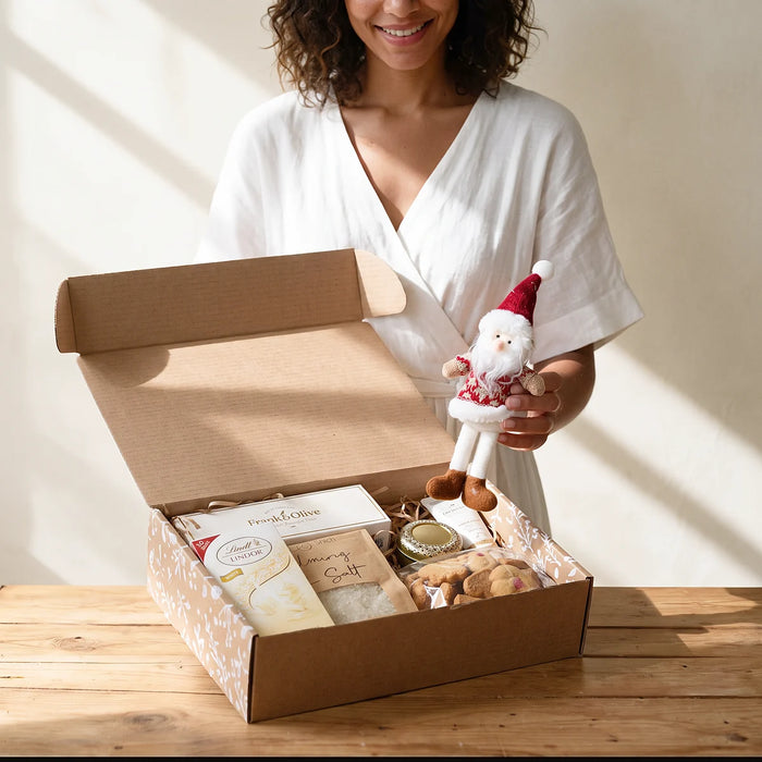 Smiling woman in a white wrap dress lifting a plush Santa out of a Christmas treat and pamper box on a pine table, Fabulous Flowers South Africa