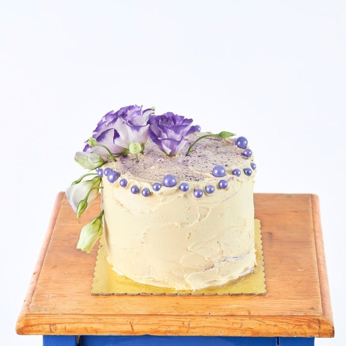 Creamy Violet Dream Cake - Fabulous Flowers & Gifts vanilla cake topped with lisianthus and purple pearls on wooden board.