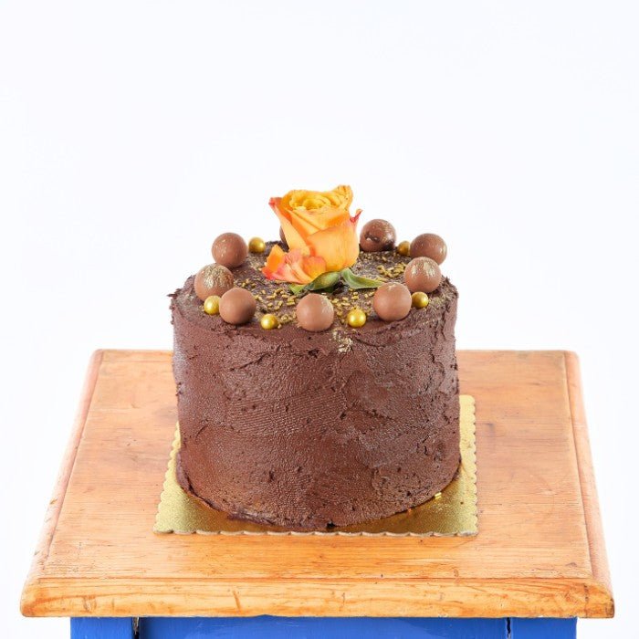 Decadent Rose Chocolate Cake topped with a vibrant orange rose and golden chocolate balls, presented on a wooden table.
