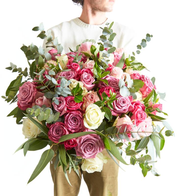 Man in white jumper holding an oversized bouquet of pink, lavender, cream, and white roses with lush greenery and eucalyptus.
