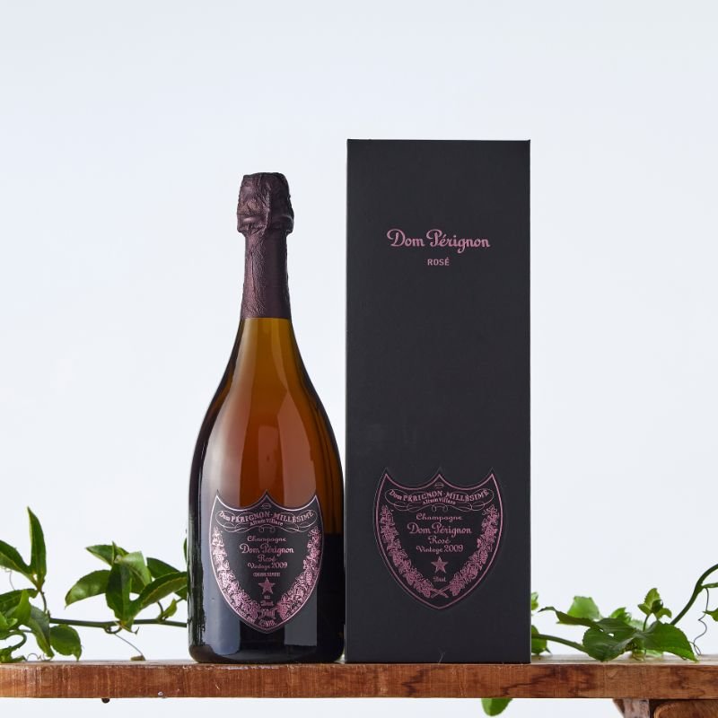 Dom Pérignon Rosé Vintage 2009 Champagne and luxury black gift box with pink branding, displayed on a wooden surface with leafy green accents.