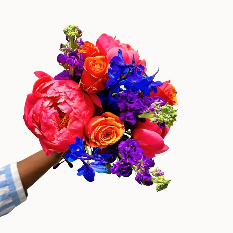 Hand holding the Electric Bloom bouquet featuring bold pink, orange, purple and blue flowers against a white background.
