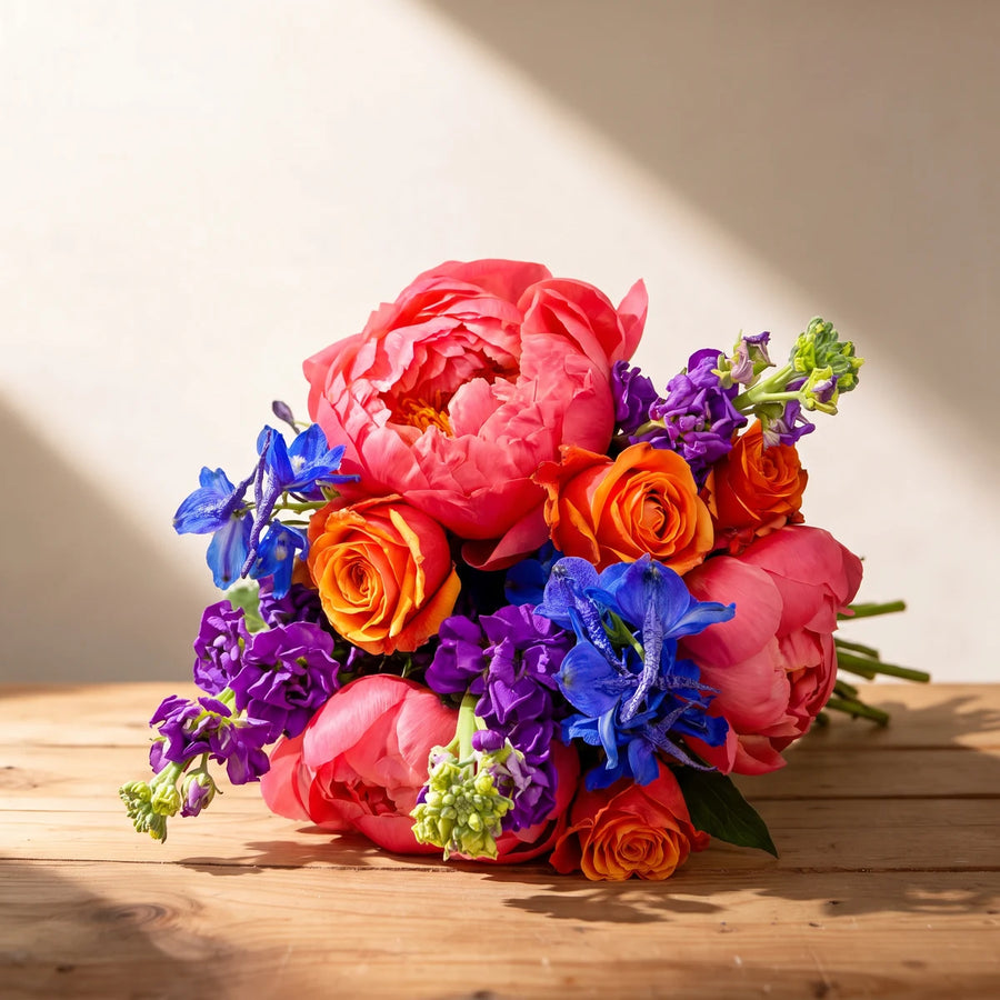 Bouquet of coral peonies, orange roses, blue delphinium and purple stocks lying on a wooden table in afternoon sun