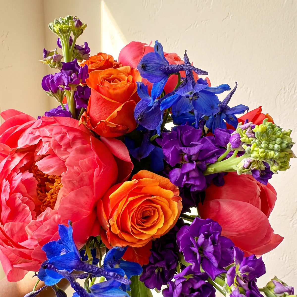 Close-up of an open coral peony with orange roses, blue delphinium and purple stocks with green buds