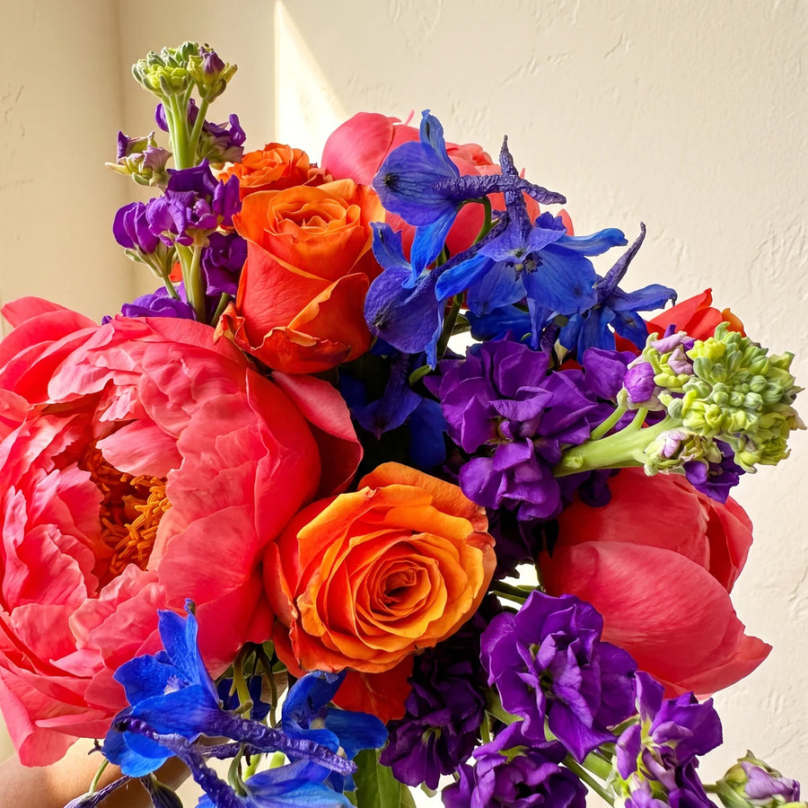 Close-up of an open coral peony with orange roses, blue delphinium and purple stocks with green buds