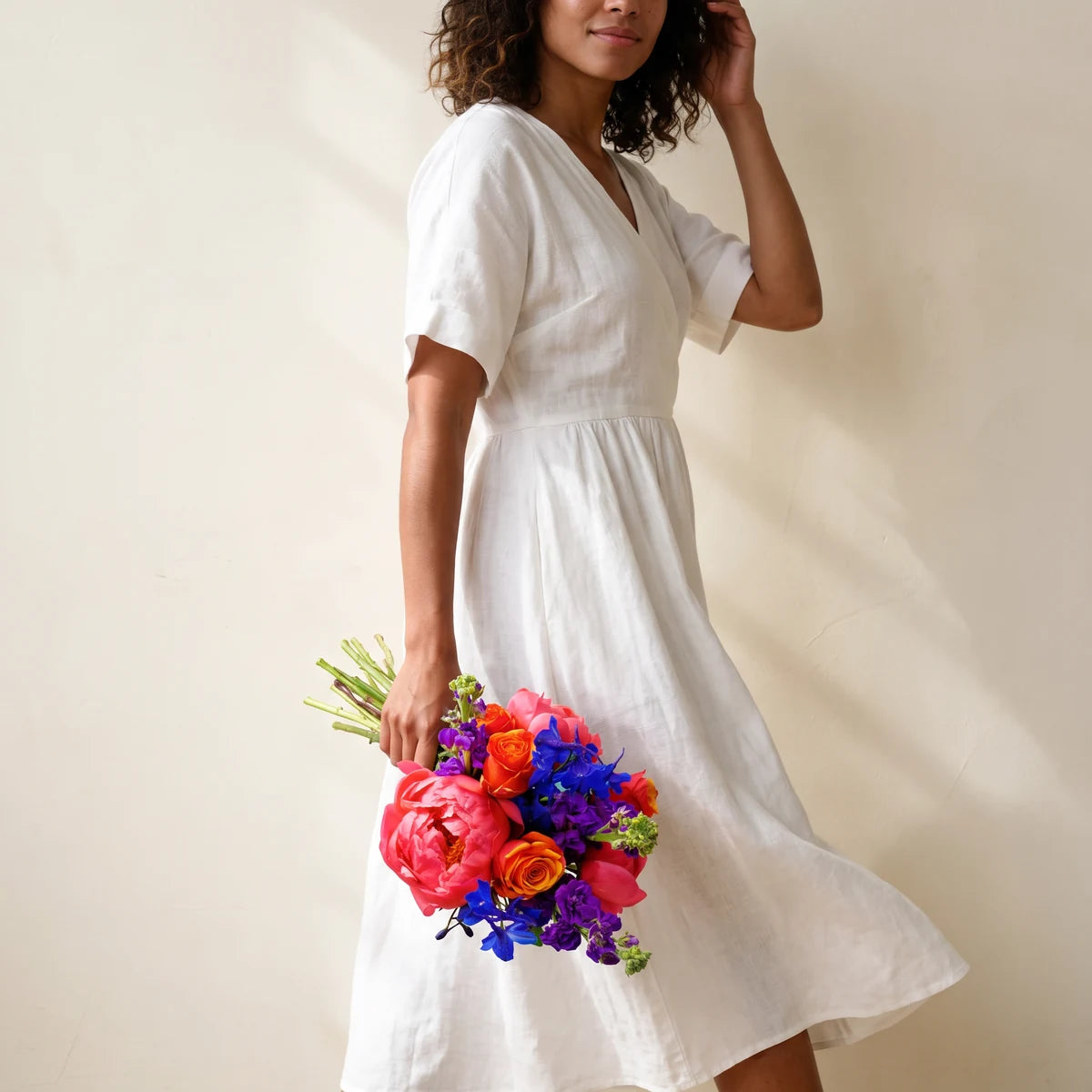 Woman with curly hair in a white dress walking with a bright bouquet of peonies and roses held at her side