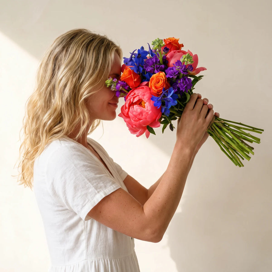 Woman with blonde hair smelling a bouquet of coral peonies, orange roses and blue delphinium